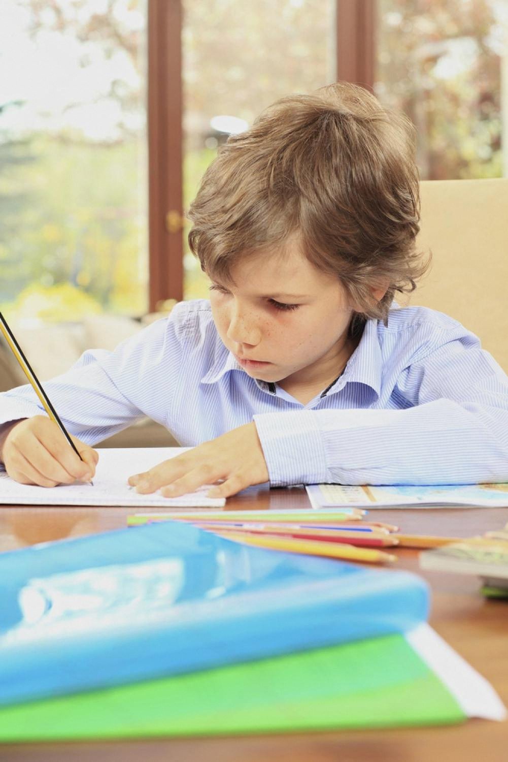 Young boy in a school shirt concentrating on his homework at a desk with colored pencils