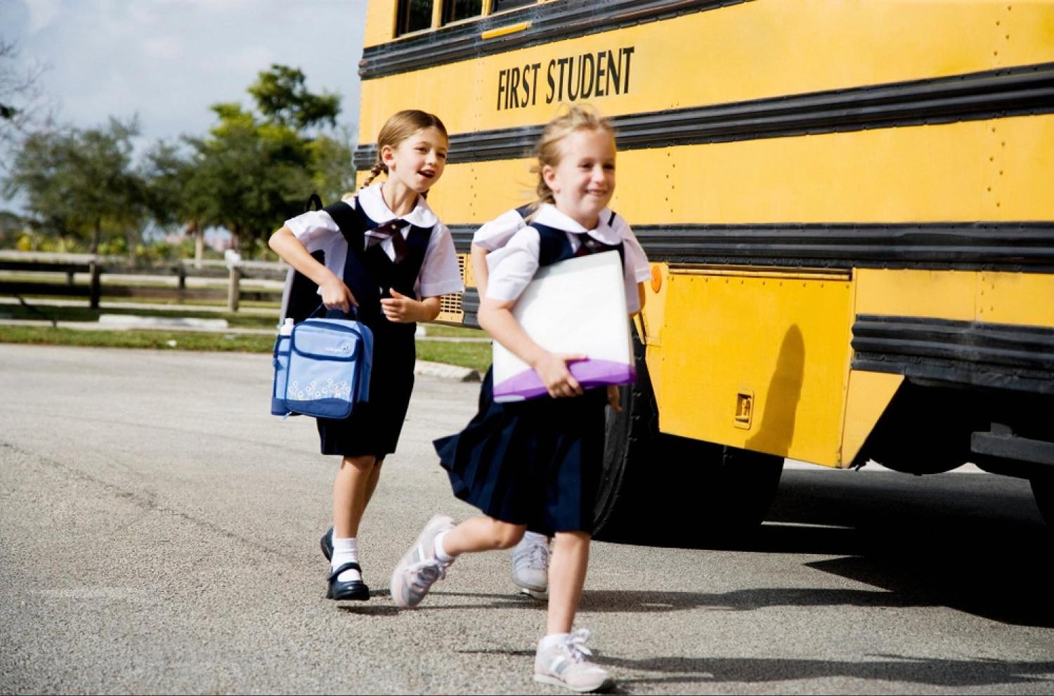 Two schoolgirls in uniform walking past a yellow school bus at the start of the school day