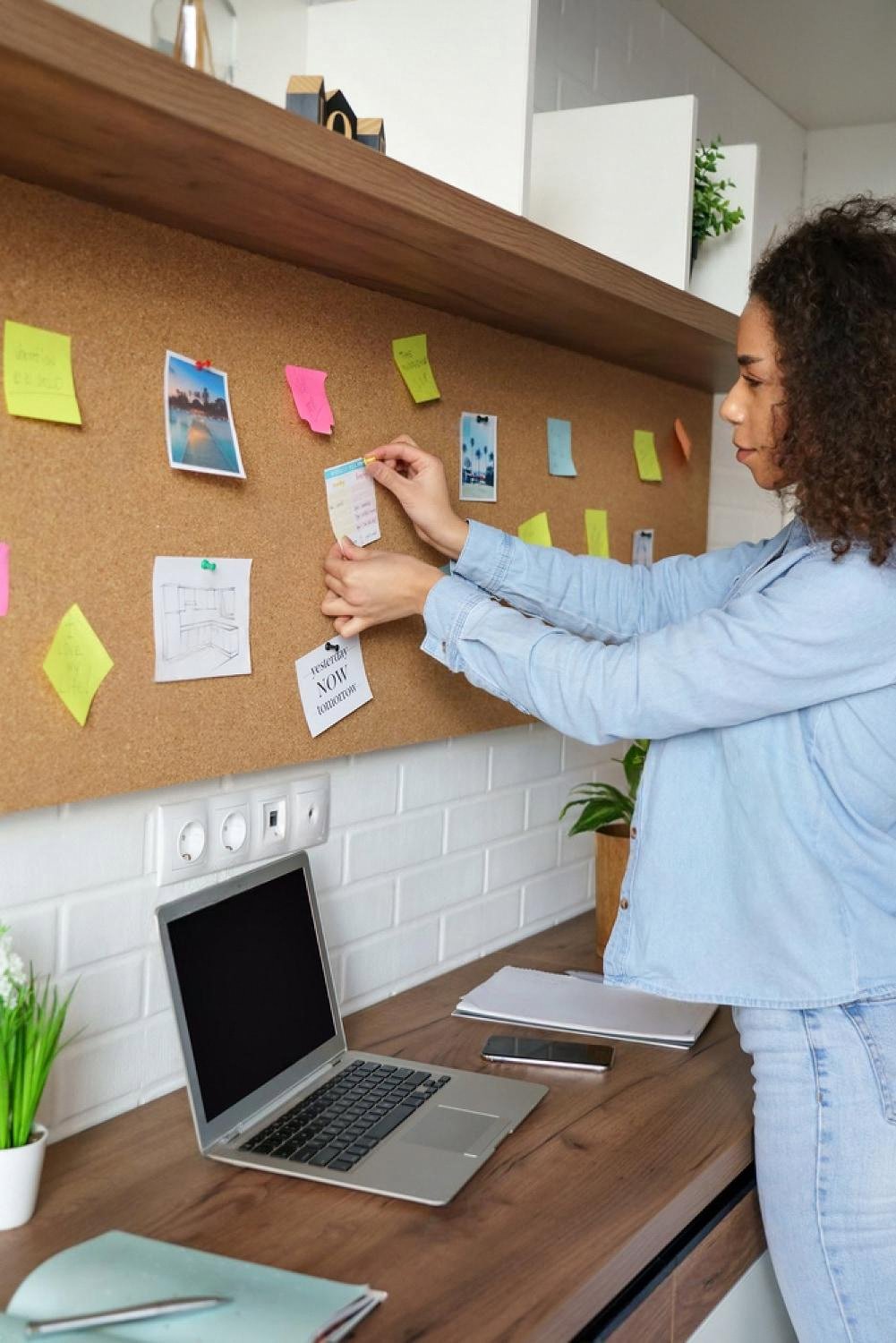 Woman pinning notes and a weekly schedule to a corkboard above a home desk